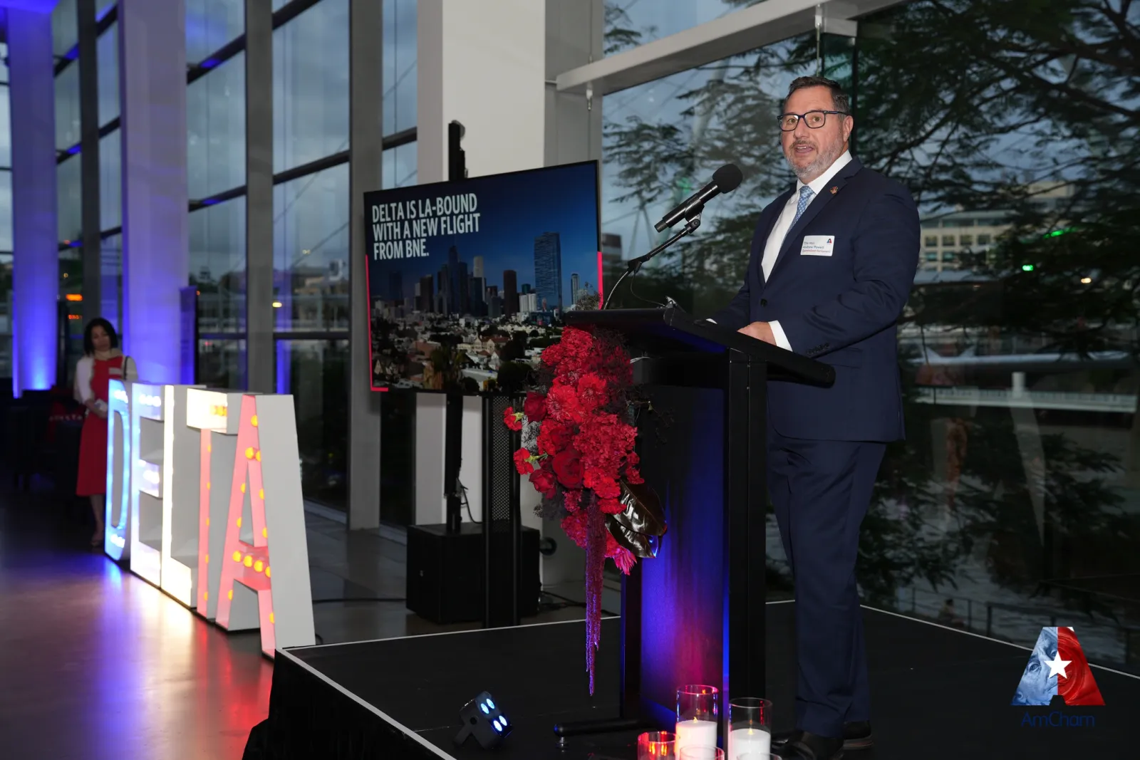 A speaker addressing delegates from the stage at an AmCham event in Brisbane, with a presentation screen and event staging behind.