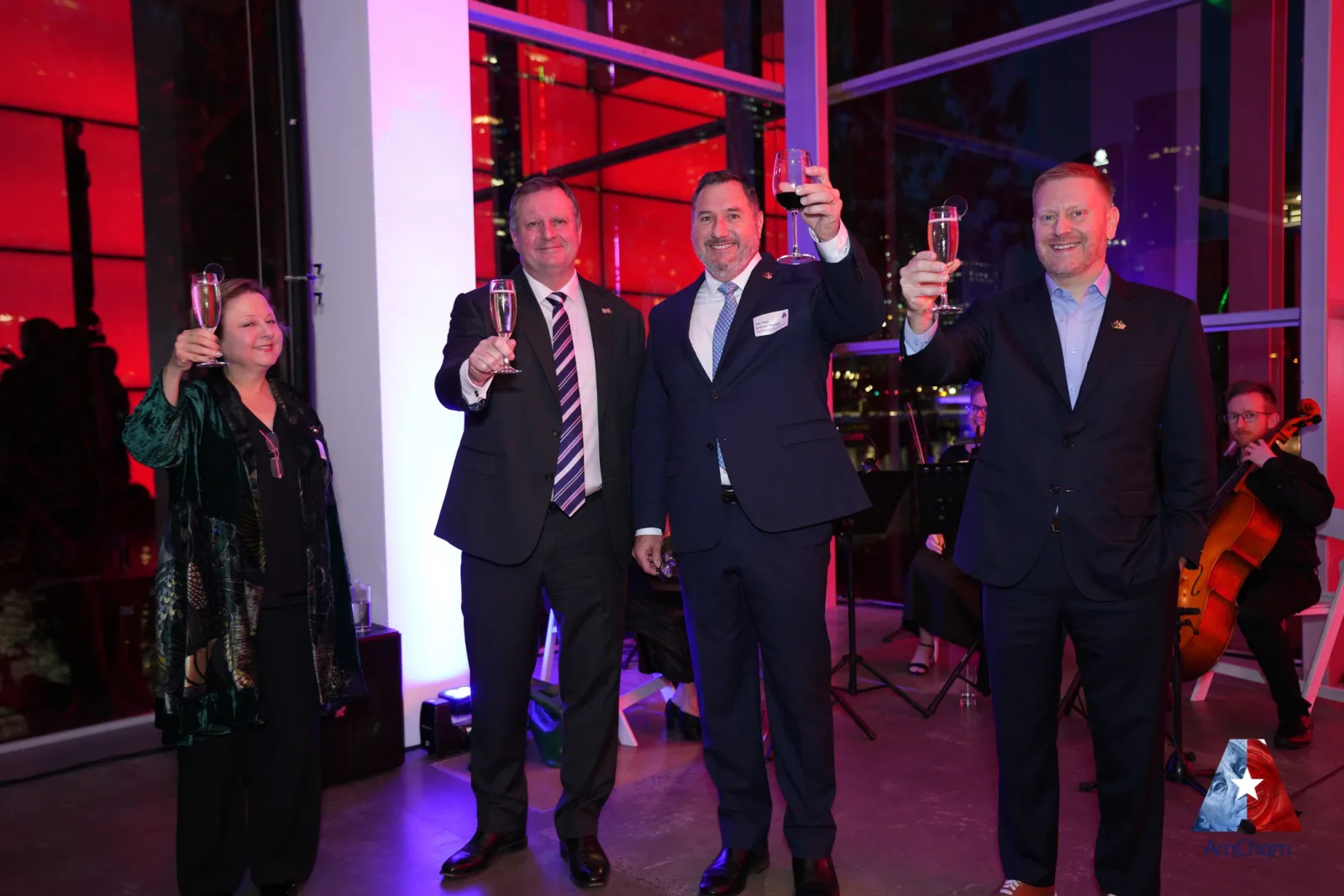 Guests raising a toast at an evening AmCham event reception, with a string musician and the Brisbane city skyline behind.